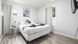 Un dormitorio con cama, mesita de noche, lámpara, un cuadro y dos ventanas con cortinas.