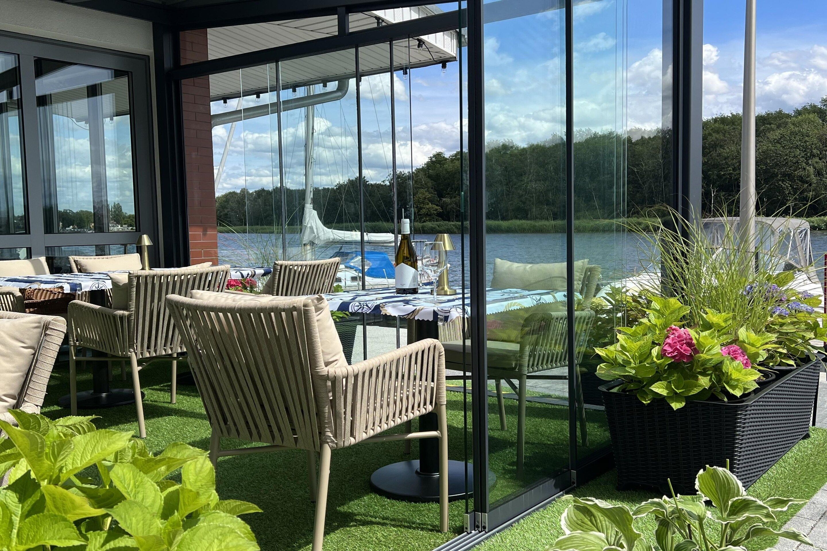 A covered outdoor seating area with wicker furniture and a table set for dining, overlooking a lake with boats.