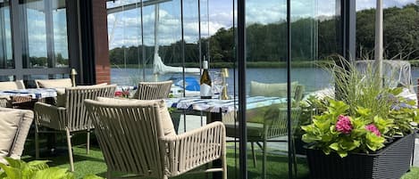 A covered outdoor seating area with wicker furniture and a table set for dining, overlooking a lake with boats.