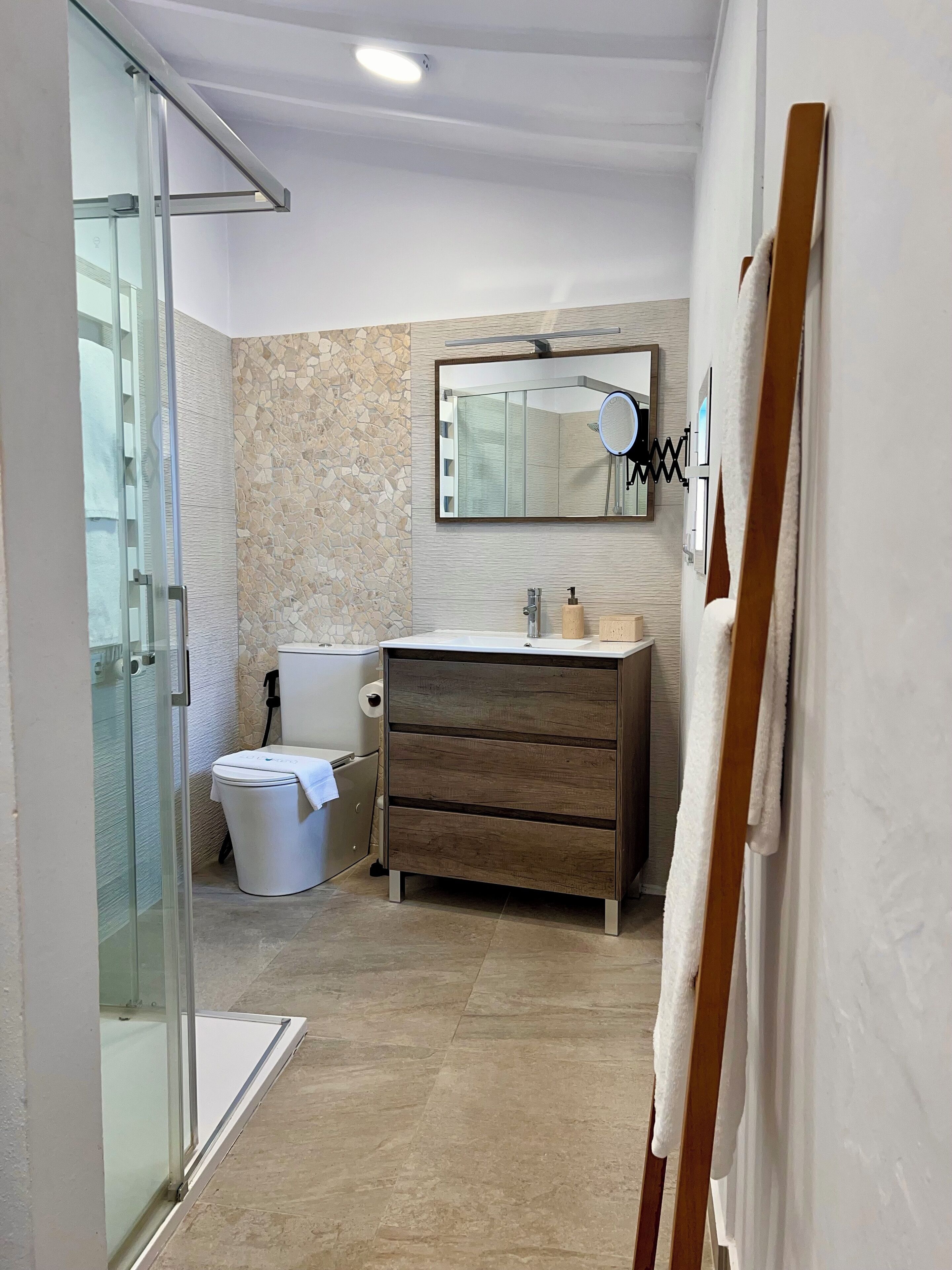 A modern bathroom with a glass shower enclosure, a white toilet, a wooden vanity with a mirror, and a towel rack.