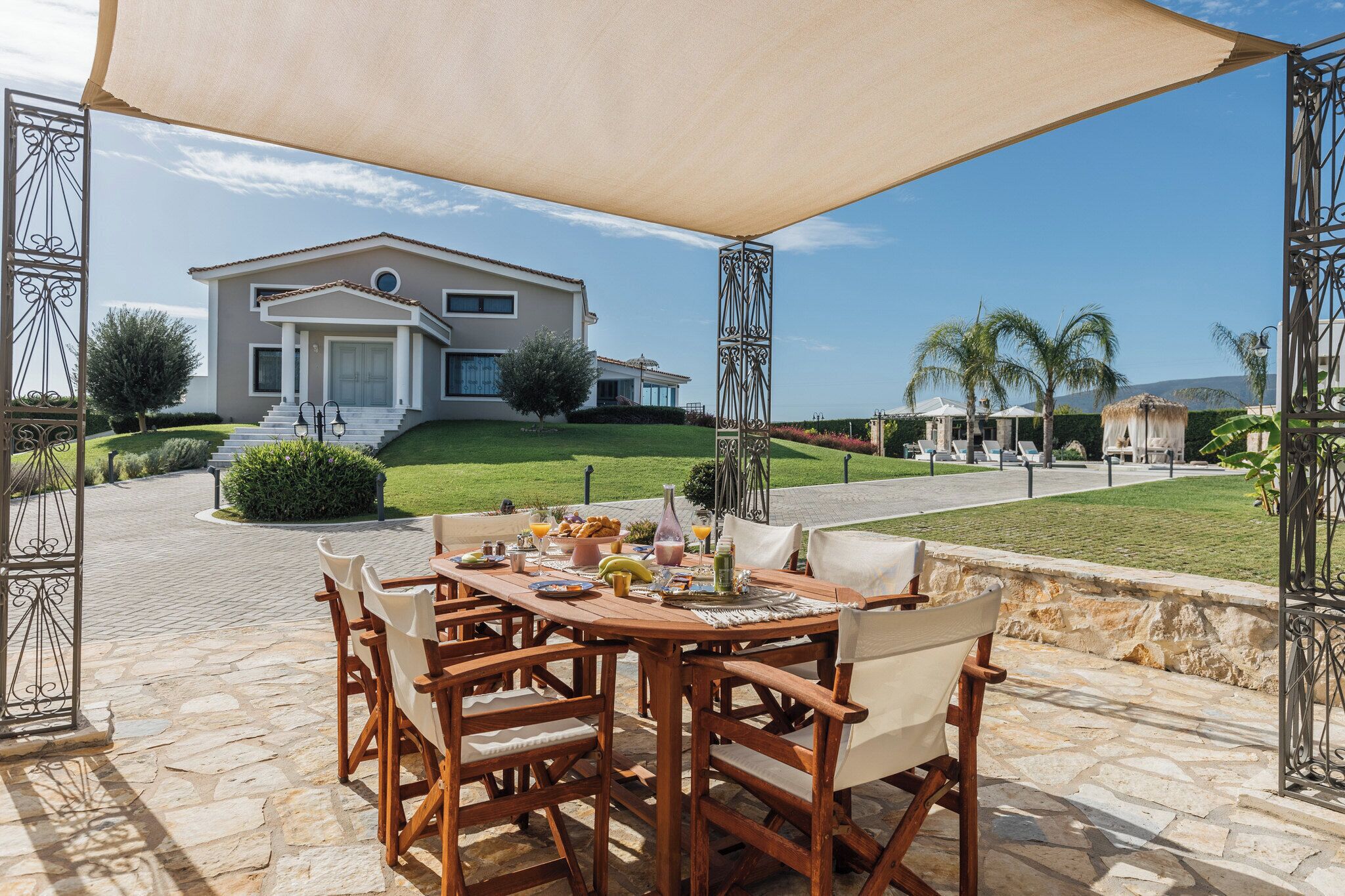 Une terrasse avec une table préparée pour le petit-déjeuner, une grande maison en arrière-plan et un ciel dégagé.