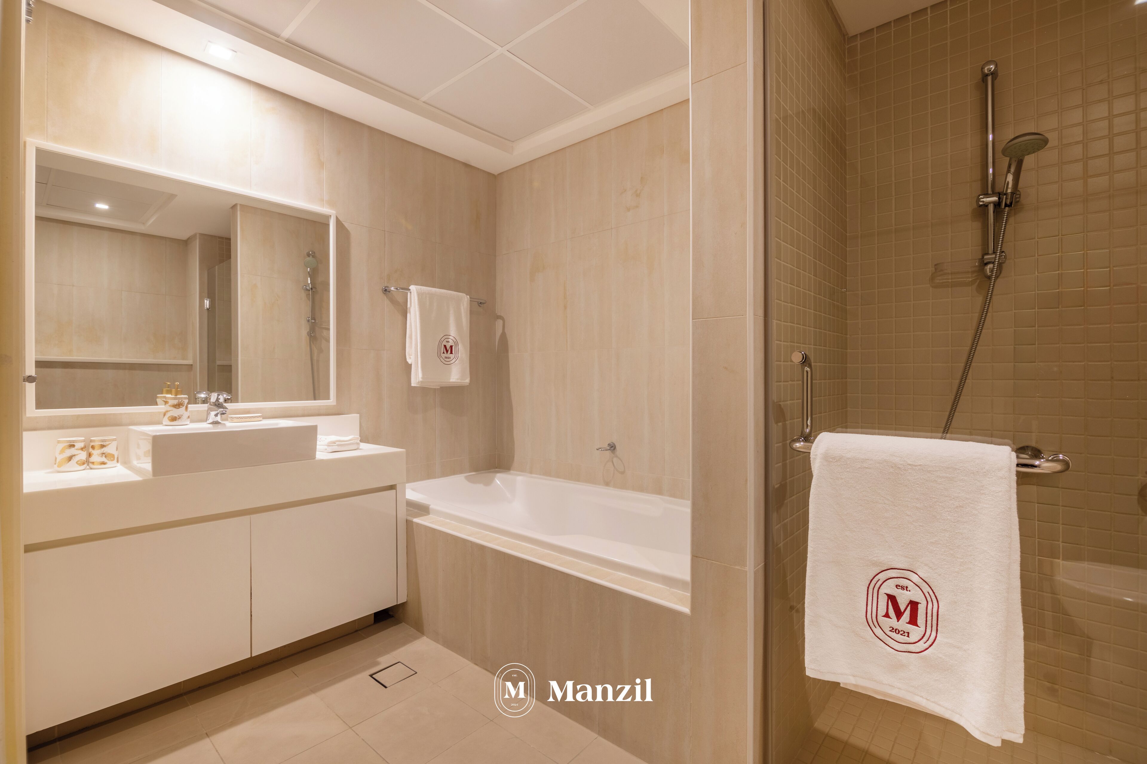 A modern bathroom with a bathtub, a shower, and a large mirror. There is a towel with a logo hanging on the wall.