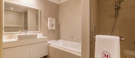 A modern bathroom with a bathtub, a shower, and a large mirror. There is a towel with a logo hanging on the wall.