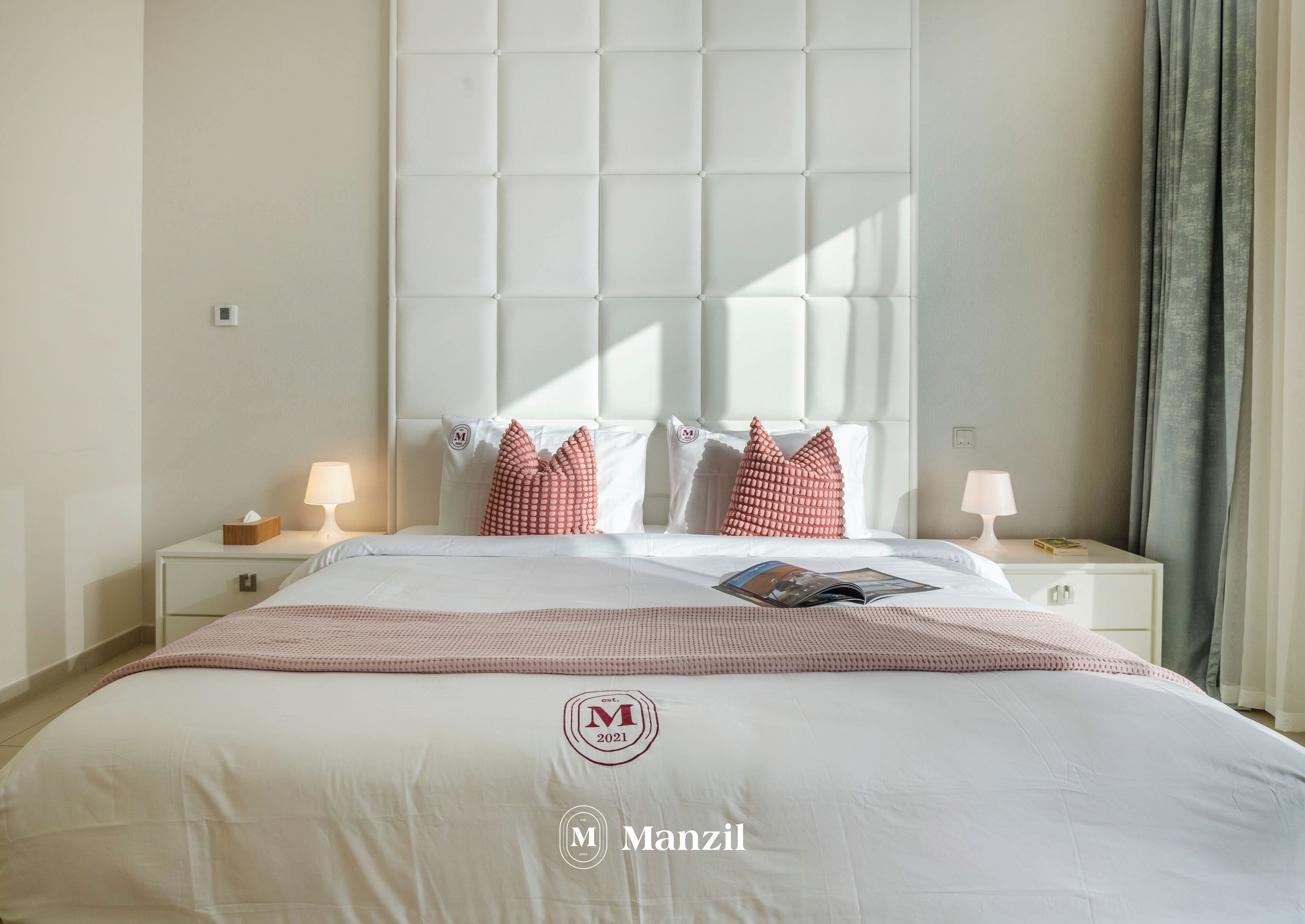 A hotel room with a large bed, two nightstands, a window with curtains, and a logo on the bedspread.