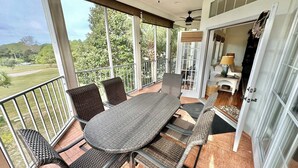 A covered outdoor patio with a table and chairs, a ceiling fan, and a view of a green landscape.