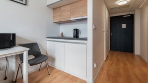 Cocina compacta con armarios blancos, encimera negra y cafetera. Hay una pequeña mesa con una silla, un cuadro en la pared y una puerta con el símbolo de Wi-Fi.