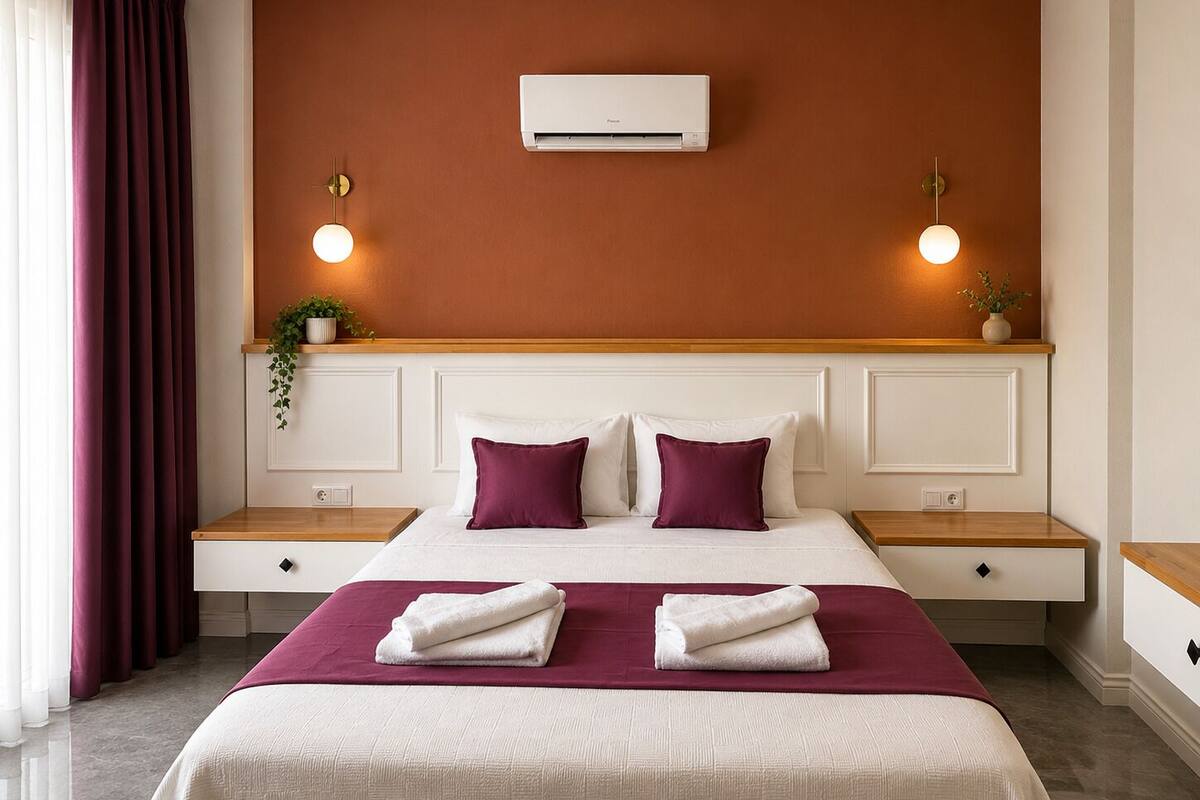 A hotel room with a bed, two bedside tables, an air conditioning unit, and a wall-mounted light fixture.