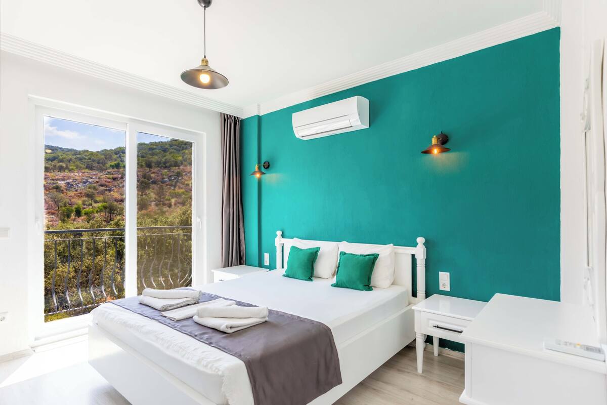 A bedroom with a large bed, a nightstand, a white dresser, an air conditioning unit, and a balcony with a view of the countryside.