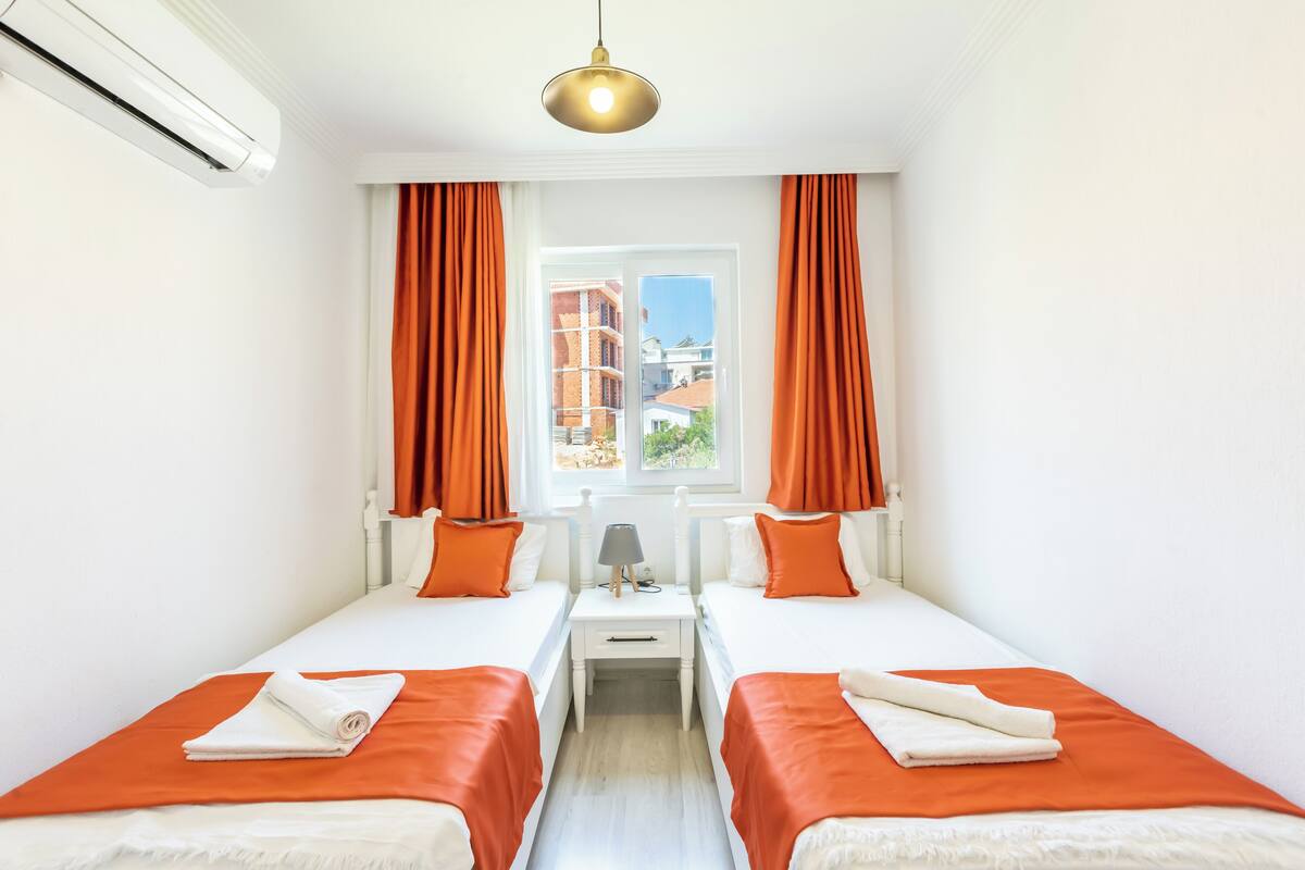 A hotel room with two beds, a window with curtains, an air conditioning unit, and a lamp.
