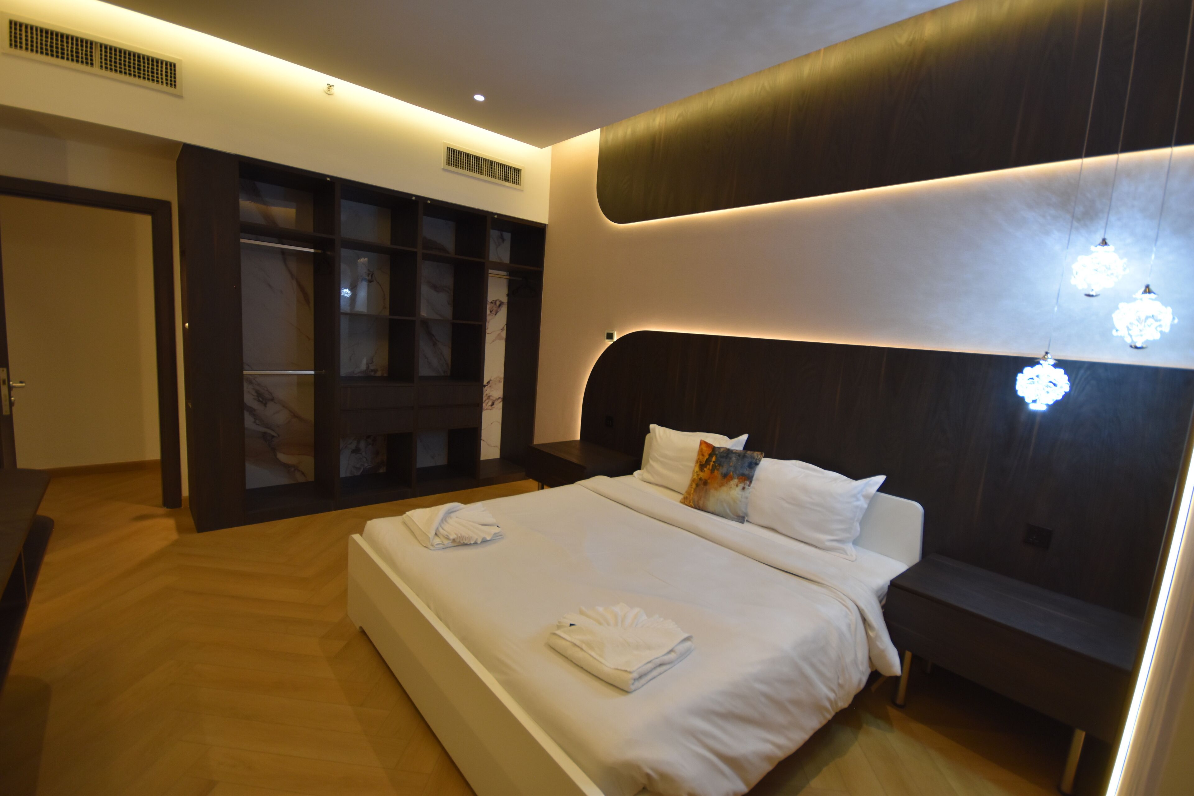 A modern hotel room with a large bed, bedside tables, a wardrobe, and a built-in bench.