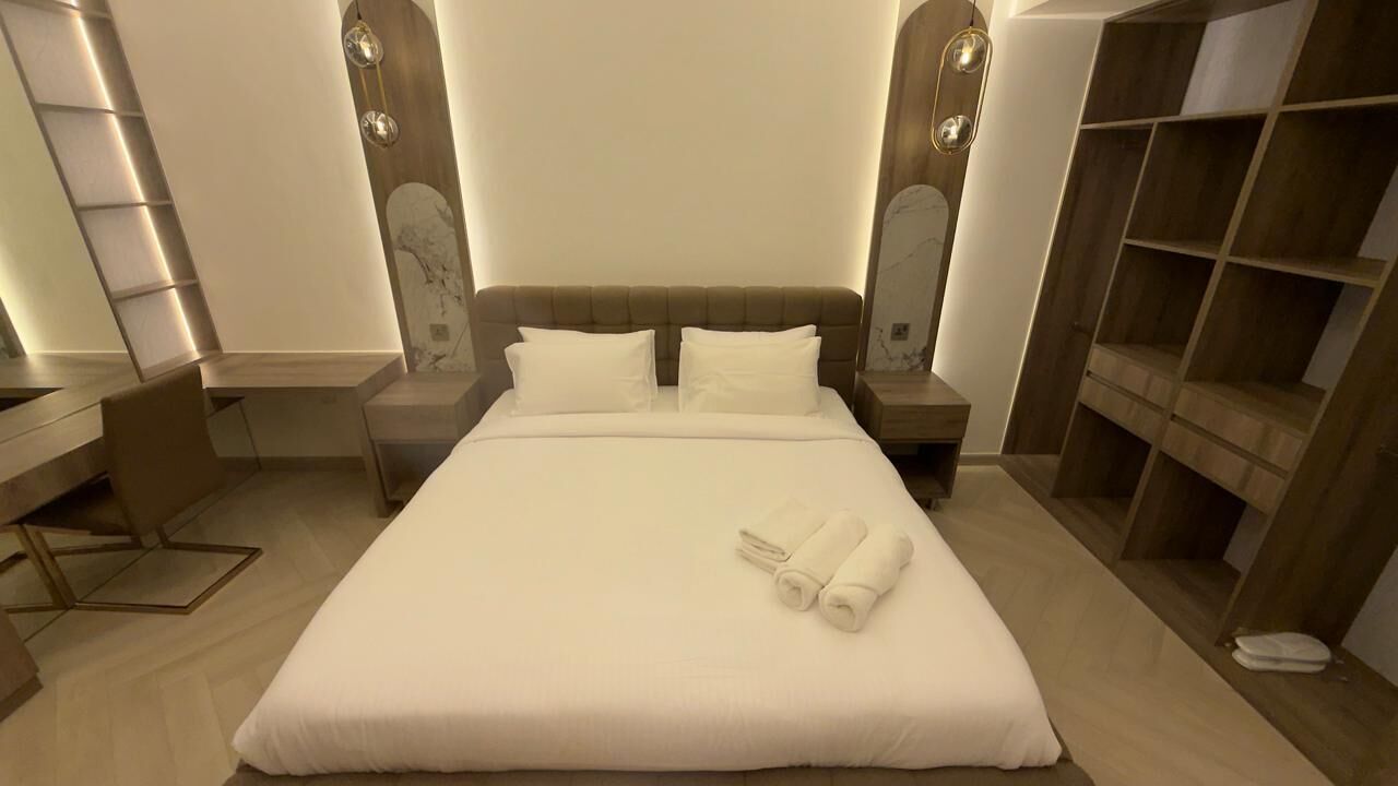 A neatly made bed with white linens and two rolled towels, a wooden nightstand with a lamp, and a built-in wooden shelving unit to the right.