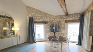 A room with a stone wall, a round dining table with chairs, a mirror with lamps, and a view through a window. - Cozy apartment in the heart of Brantôme, private terrace (Brantôme en Périgord-Brantôme en Périgord)