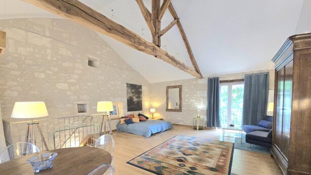 A rustic room with exposed wooden beams, a stone wall, a bed with blue bedding, a sofa, a wooden cabinet, and a dining table with chairs. - Cozy apartment in the heart of Brantôme, private terrace (Brantôme en Périgord-Brantôme en Périgord)