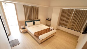 A hotel room with a bed, bedside tables, a small shelf with toiletries, and curtains on the windows. - EXCELLENT HOMES (Nashik)