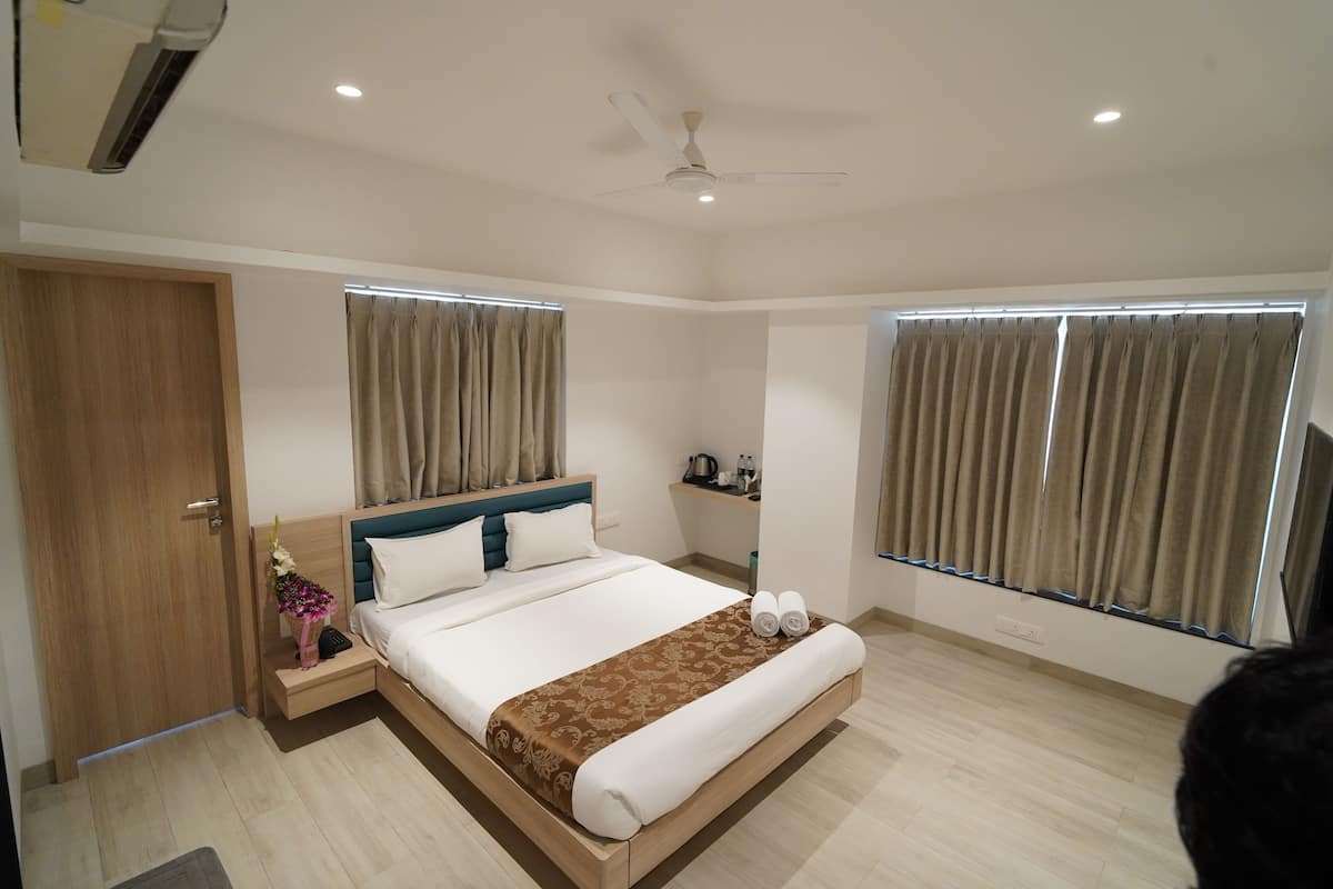 Quarto de hotel moderno com uma cama grande, mesa de cabeceira com abajur, ventilador de teto e janelas amplas com cortinas.