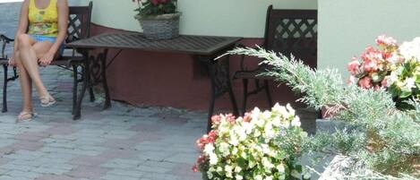 A person sitting on a bench outside a building with a table and chairs nearby. There are potted plants with flowers and shrubs around.