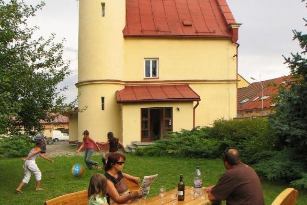 A yellow building with a red roof, a wooden table with bottles and a menu, and people outside.
