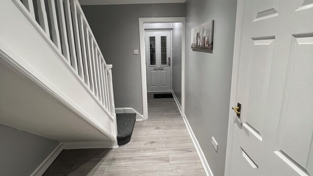 A hallway with a wooden floor, white walls, and a white door at the end. - Spacious 3 Bed Home - Ideal for Families & Groups (Linford)