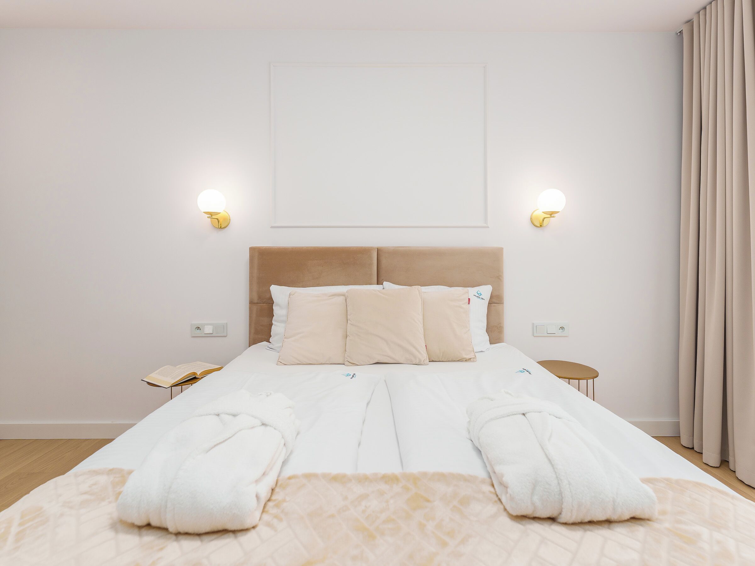 Ein ordentlich bezogenes Bett mit weißen Leinen und zwei Kissen, ein Nachttisch mit Lampe sowie eine Wandleuchte.