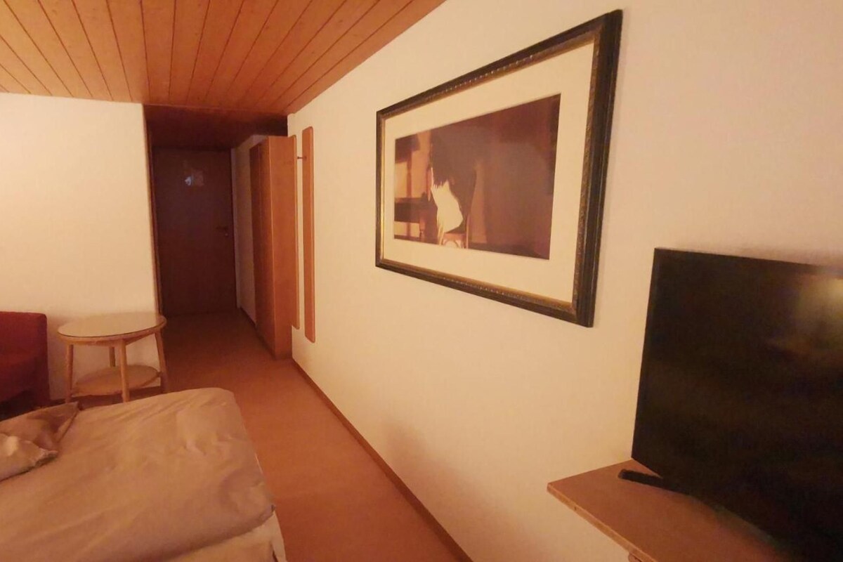 A hotel room with a bed, a red armchair, a small table, a television, and a framed picture on the wall.
