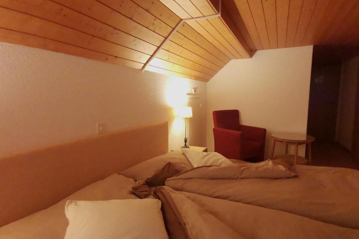 A room with a sloped ceiling, a bed with pillows, a red chair, a small table, and a lamp.