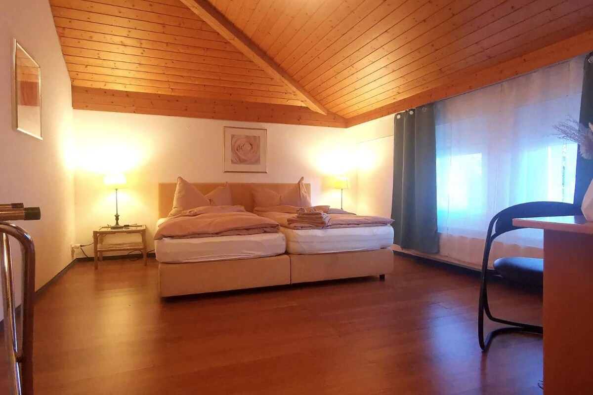 A bedroom with a wooden ceiling, a bed with pillows, a nightstand with a lamp, a chair, and a vase with flowers.