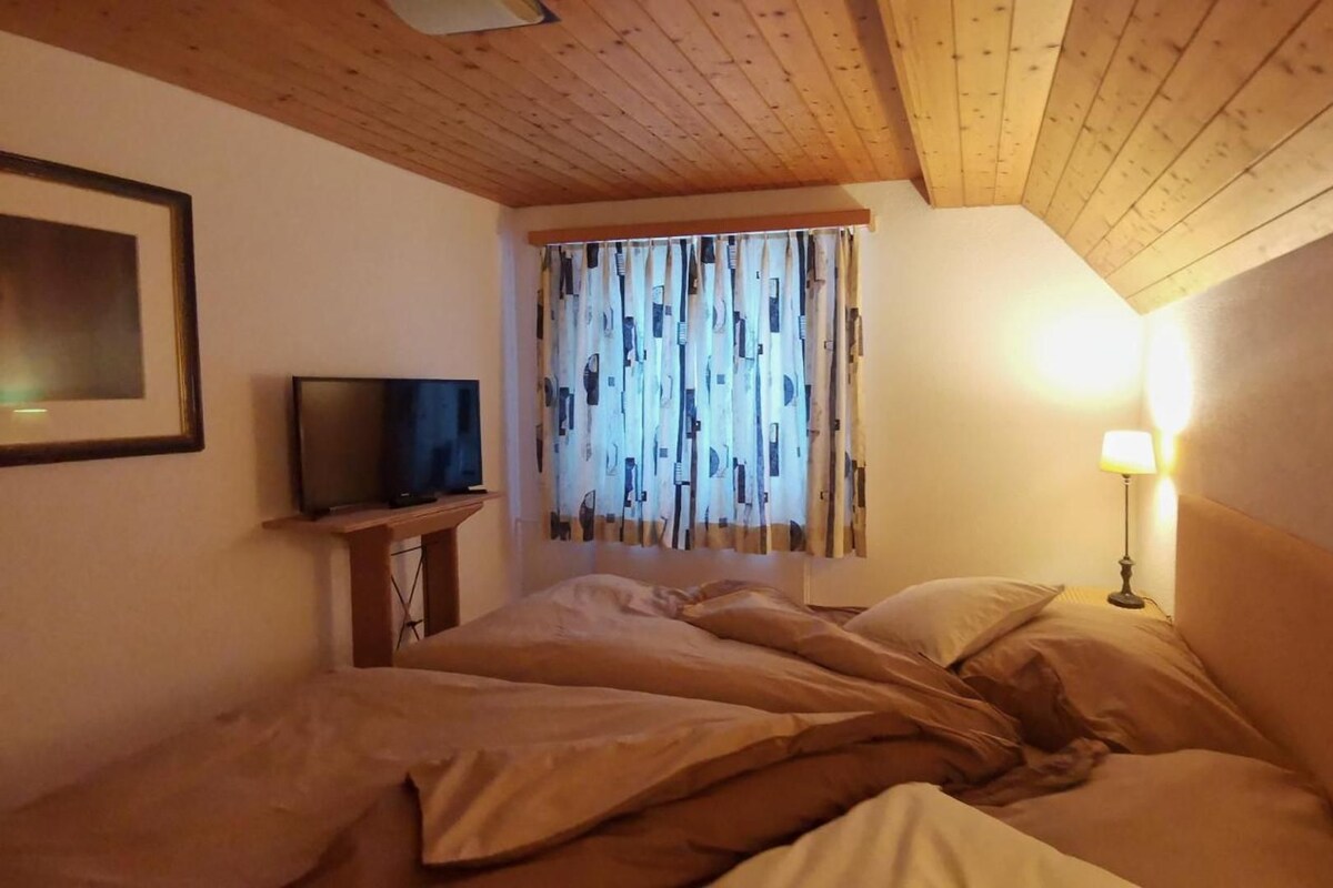 A bedroom with a bed, a television on a stand, a window with patterned curtains, and a bedside lamp.