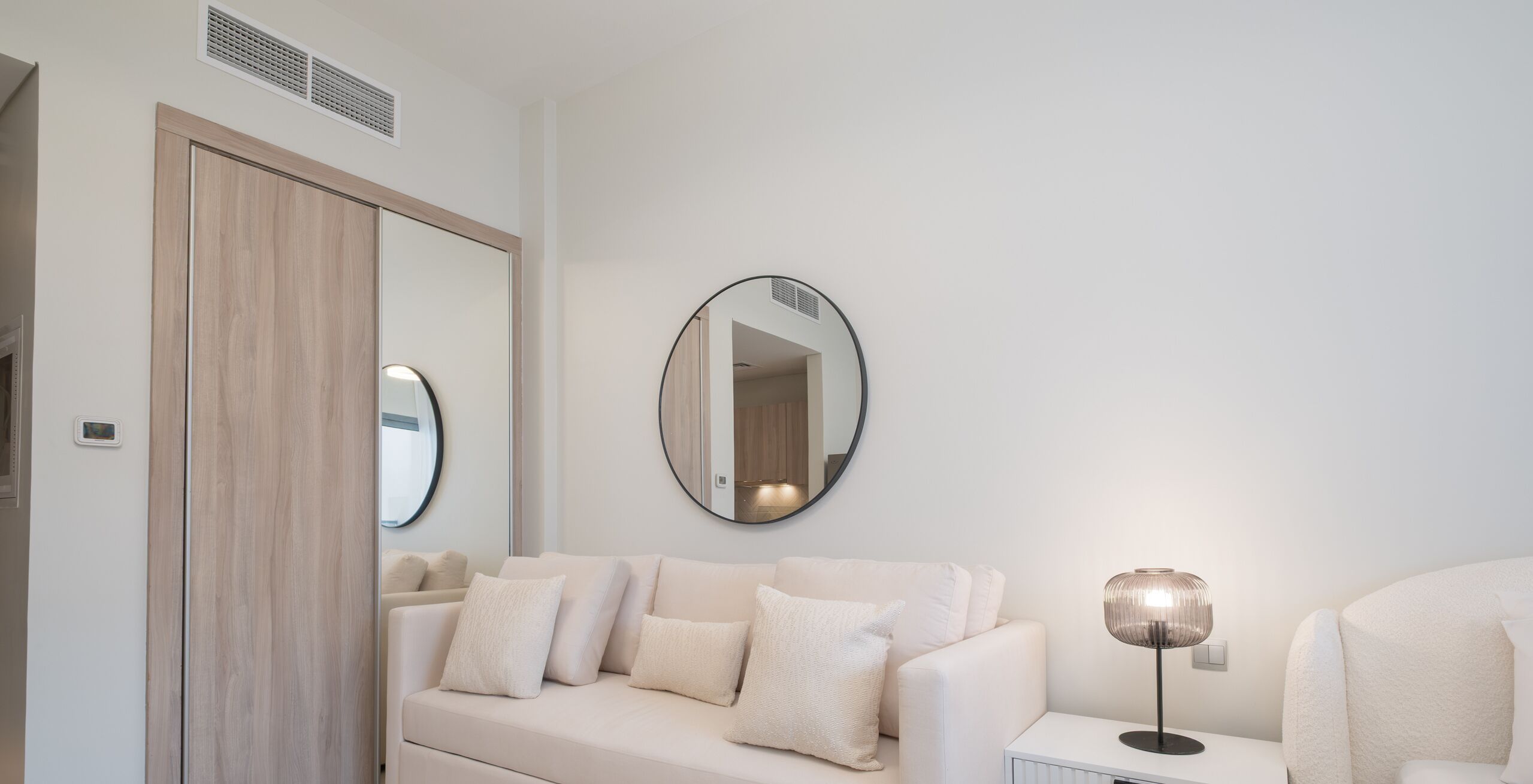 A modern bedroom with a white sofa, a round mirror, and a bedside table with a lamp.