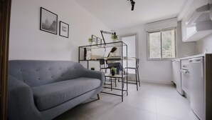 Een moderne keuken met witte apparaten, een grijze bank en een persoon achter een bureau met een plant.