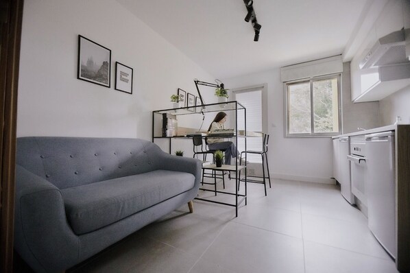 A modern kitchen with white appliances, a grey sofa, and a person working at a desk with a plant. - Hostel just 2 minutes from the beach! (Perillo)