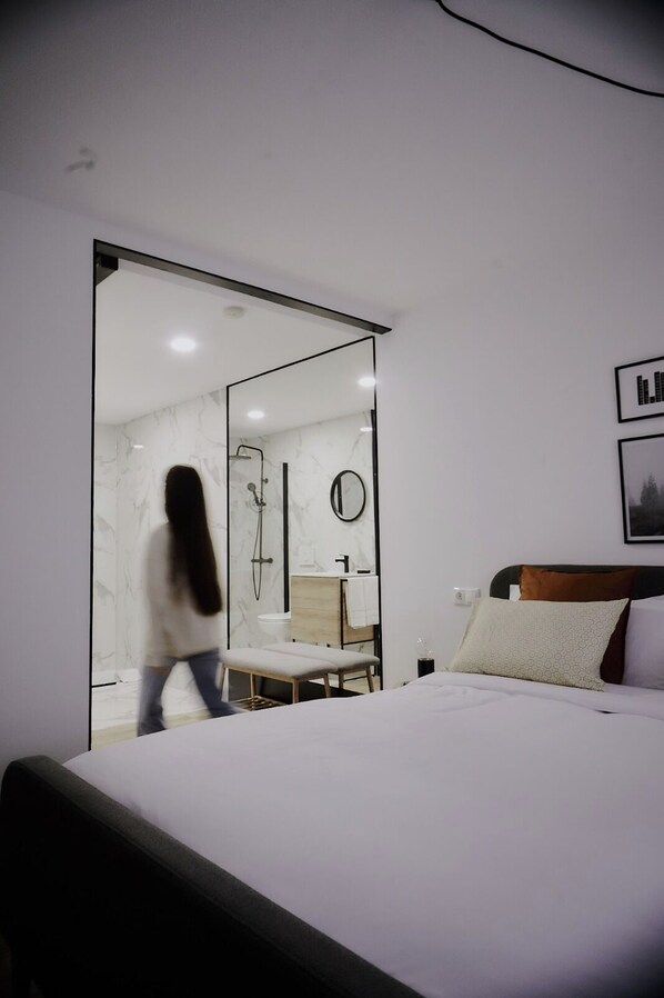 A bedroom with a large bed, a bedside table, a mirror, and a view of a bathroom with a shower and toilet.