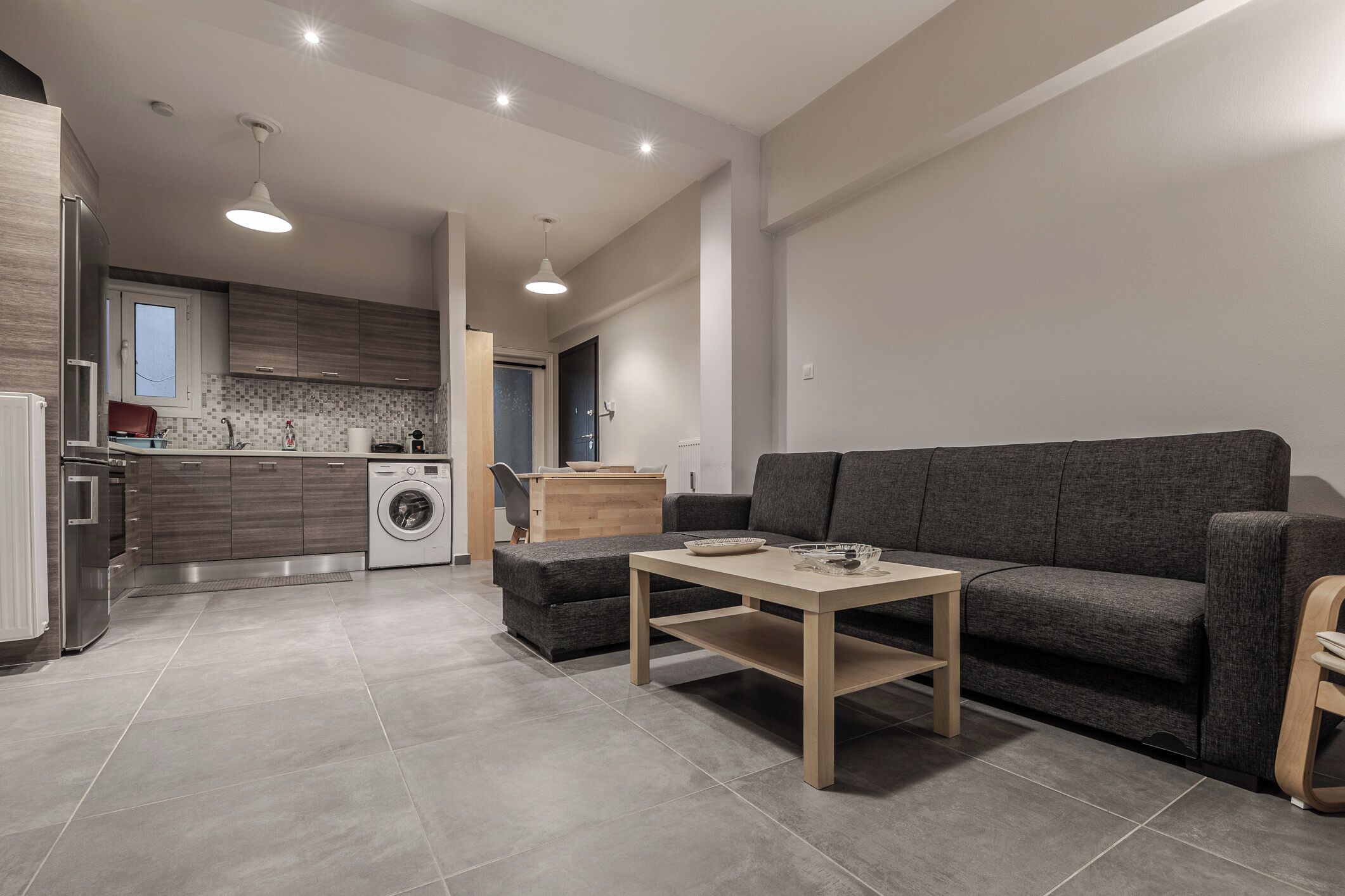 Una cocina moderna con lavadora, un sofá gris y una mesa de madera.