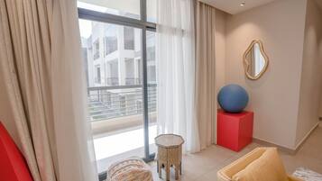 A modern hotel room with a large window, a round woven ottoman, a hexagonal side table, a red cube, and a blue exercise ball on a red pedestal.