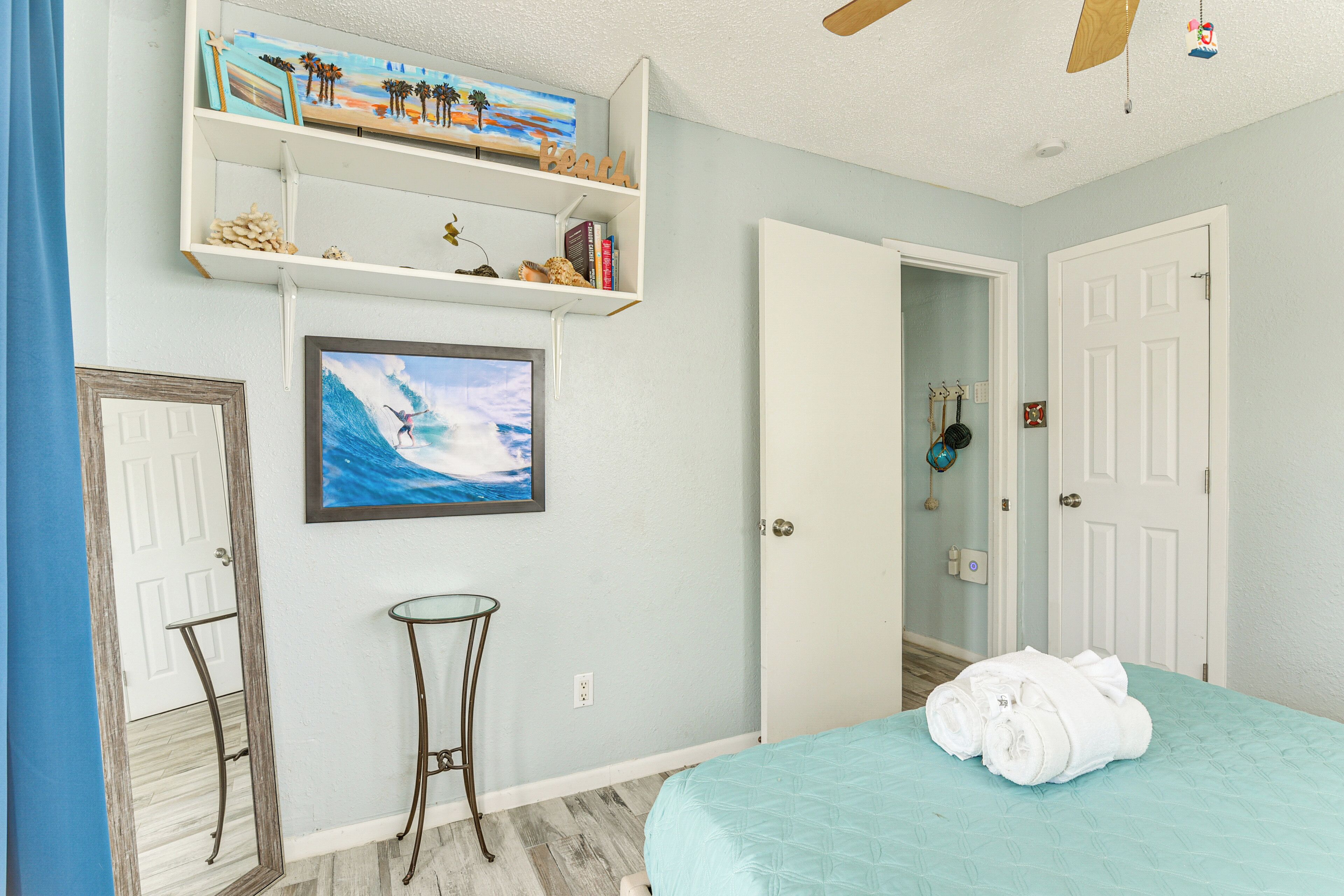 A bedroom with a bed, a small table, a shelf with books and decorative items, a painting of a surfer, and a door.