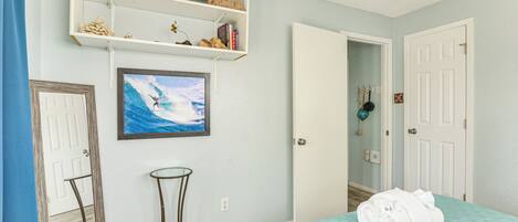 A bedroom with a bed, a small table, a shelf with books and decorative items, a painting of a surfer, and a door.
