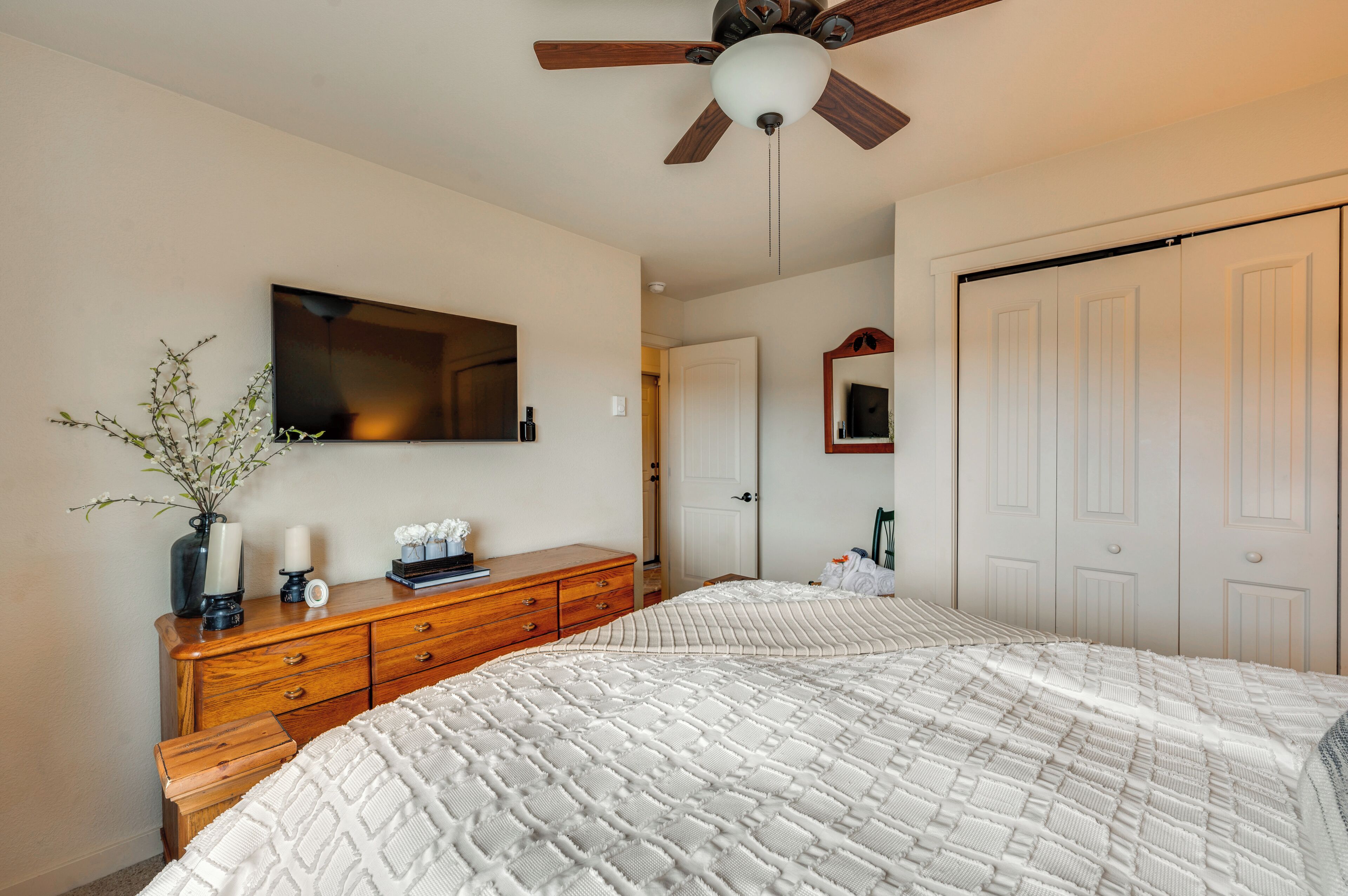 A bedroom with a bed, wooden dresser, vanity, mirror, ceiling fan, and closet.