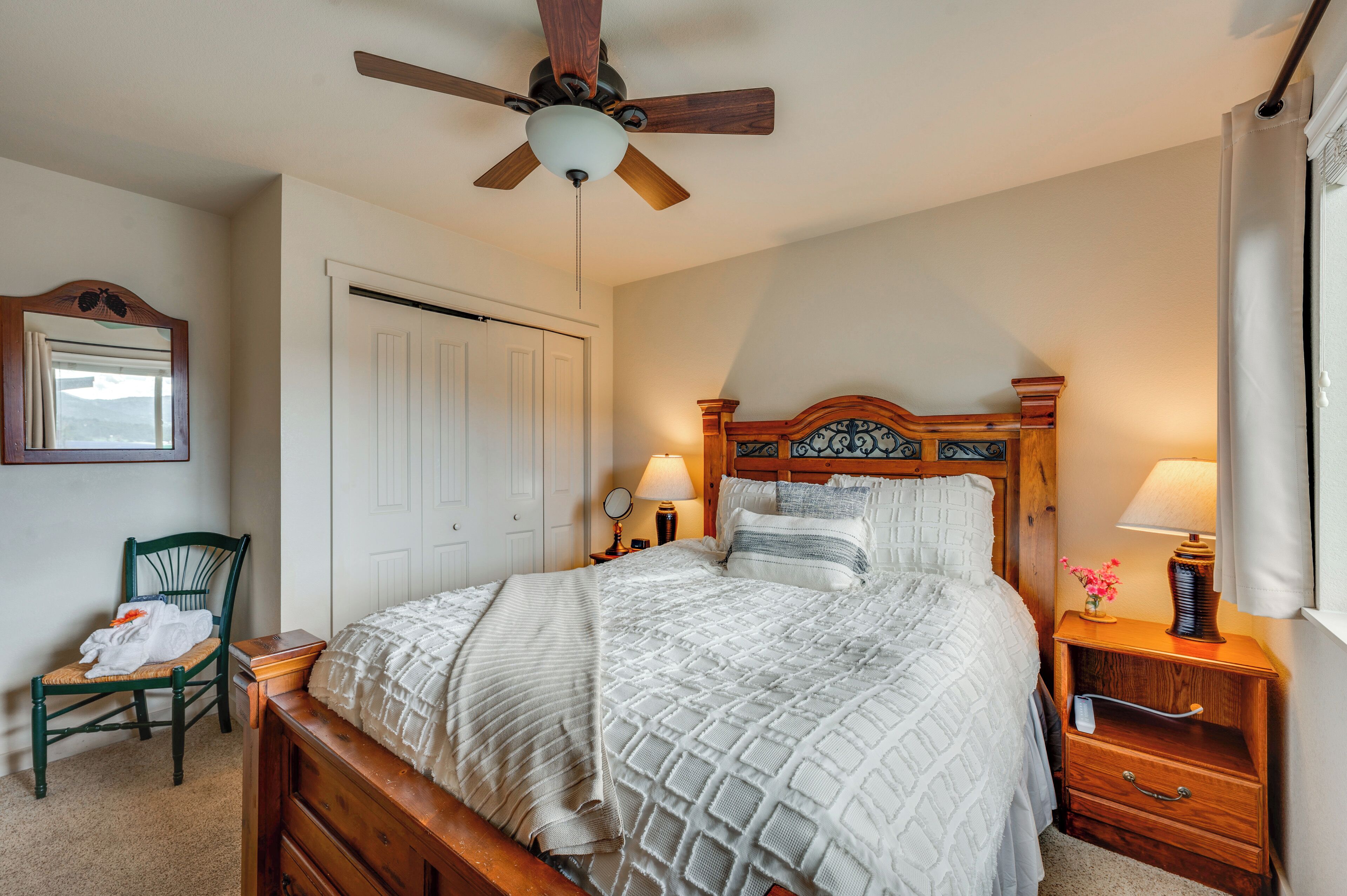 A bedroom with a wooden bed, a ceiling fan, a nightstand with a lamp, a chair, and a door.