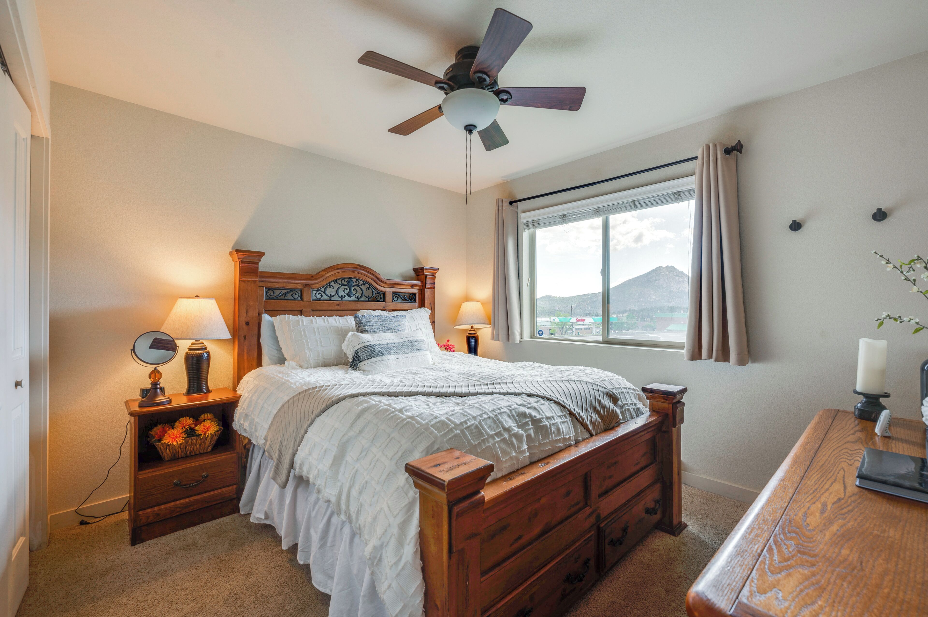 A bedroom with a wooden bed, two bedside tables with lamps, a ceiling fan, a window with curtains, and a view of a mountain.