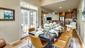 Un comedor con una mesa larga dispuesta para seis personas, rodeada de sillas, con vista a la cocina al fondo.