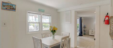 A dining area with a white table and chairs, a vase with flowers, and a window with blinds.
