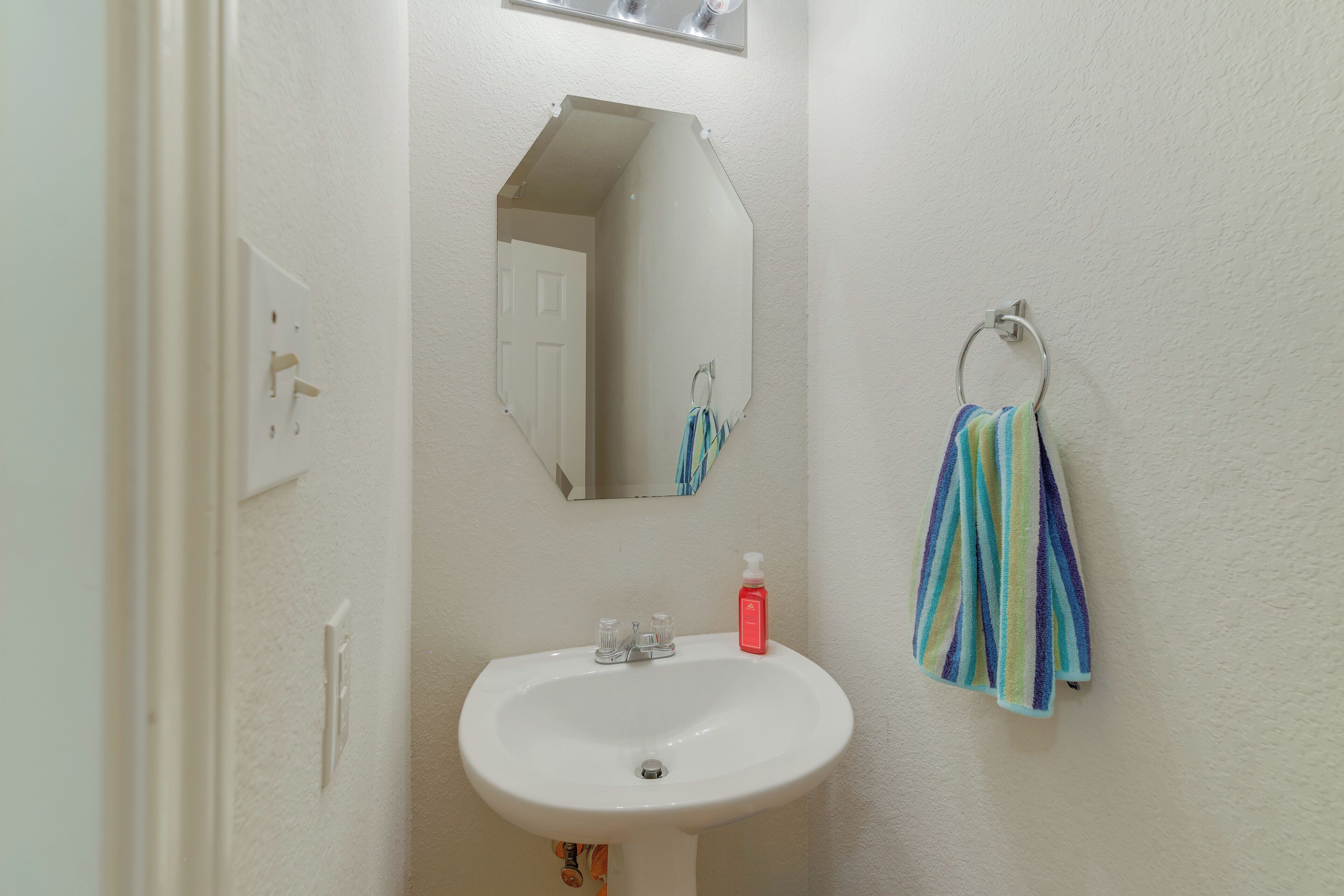 Un baño con un lavabo blanco, un espejo, un perchero con una toalla a rayas y una botella roja sobre el lavabo.
