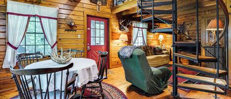 A cozy cabin interior with a dining area, a green armchair, a staircase, and a red door.
