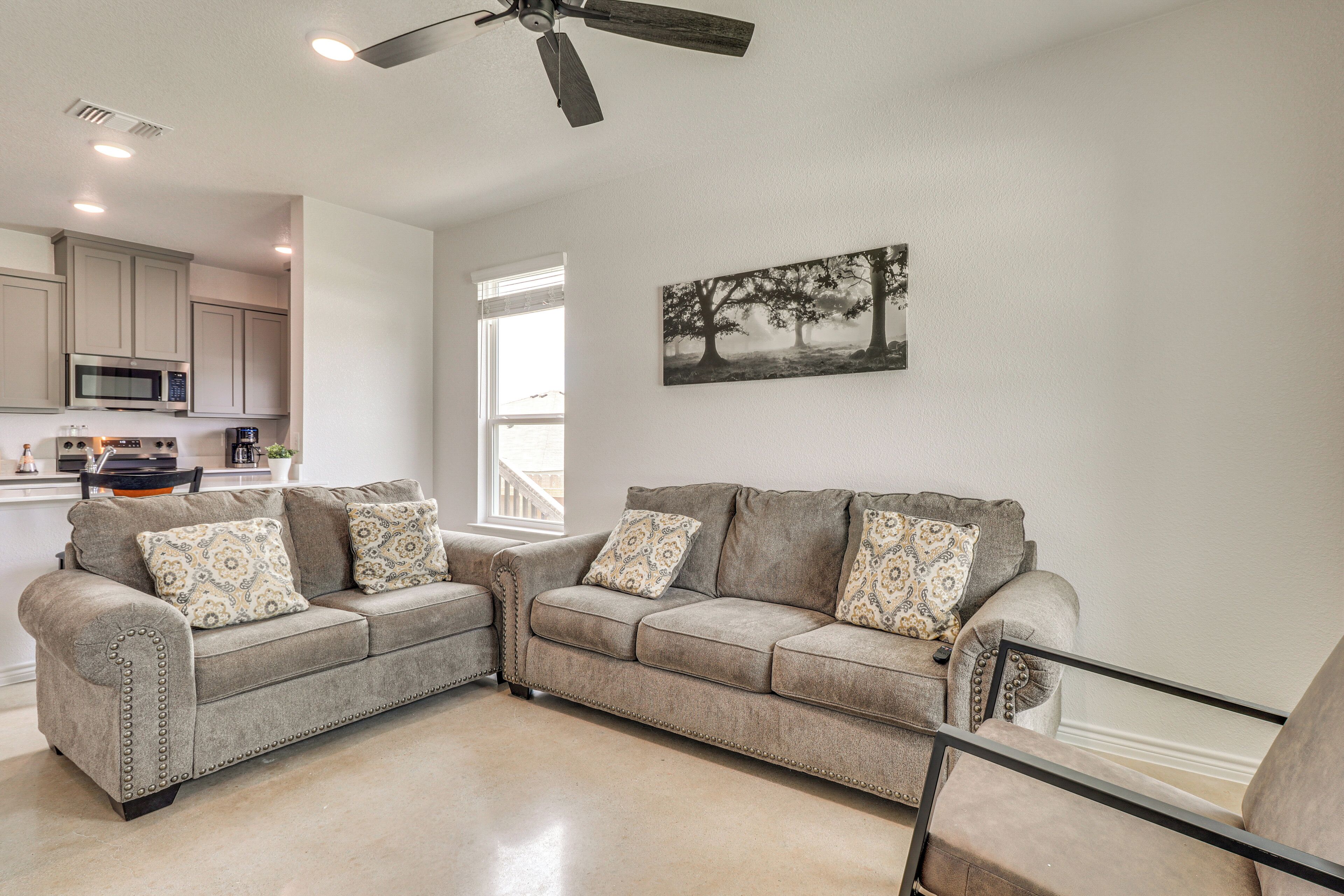 A living room with two grey sofas, a coffee table, and a ceiling fan. There is a kitchen in the background with white cabinets and a microwave.