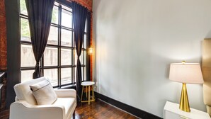 Ein Zimmer mit einem weißen Sofa, einem kleinen runden Tisch, einer goldfarbenen Lampe und einem Fenster mit schwarzen Vorhängen.