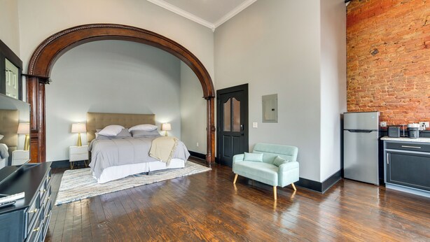 Ein Schlafzimmer mit großem Bogenfenster, einem Bett, Nachttischlampen, einem hellblauen Sessel und einer Küchenzeile mit einem Kühlschrank.