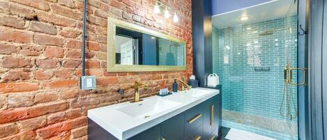 A modern bathroom with a brick wall, a large mirror, dual sinks, and a shower area.
