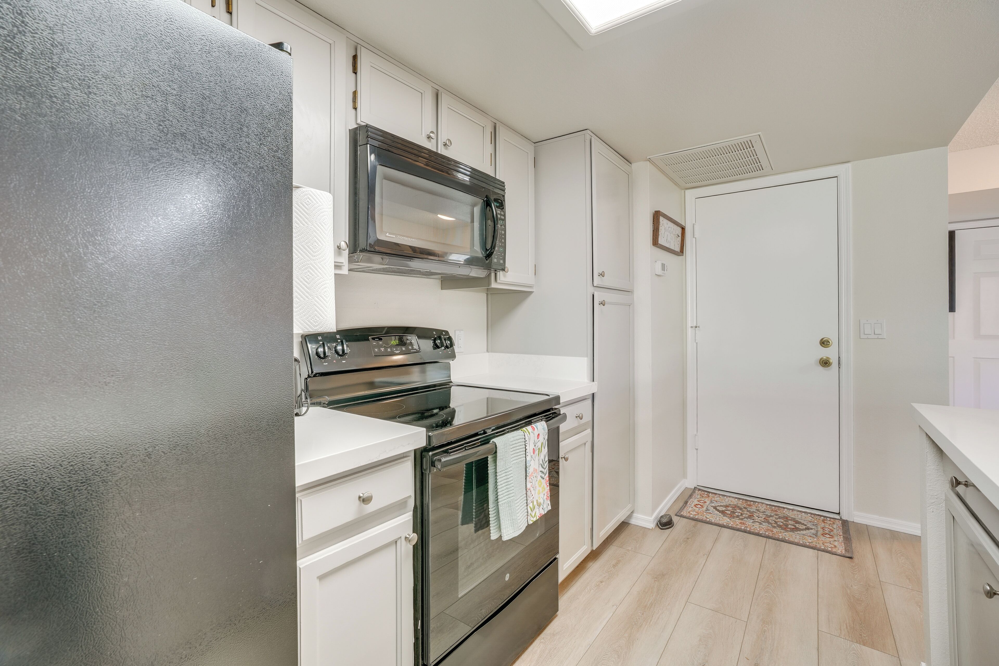 Una cocina con armarios blancos, un refrigerador de acero inoxidable, un microondas, un horno y una puerta con felpudo.