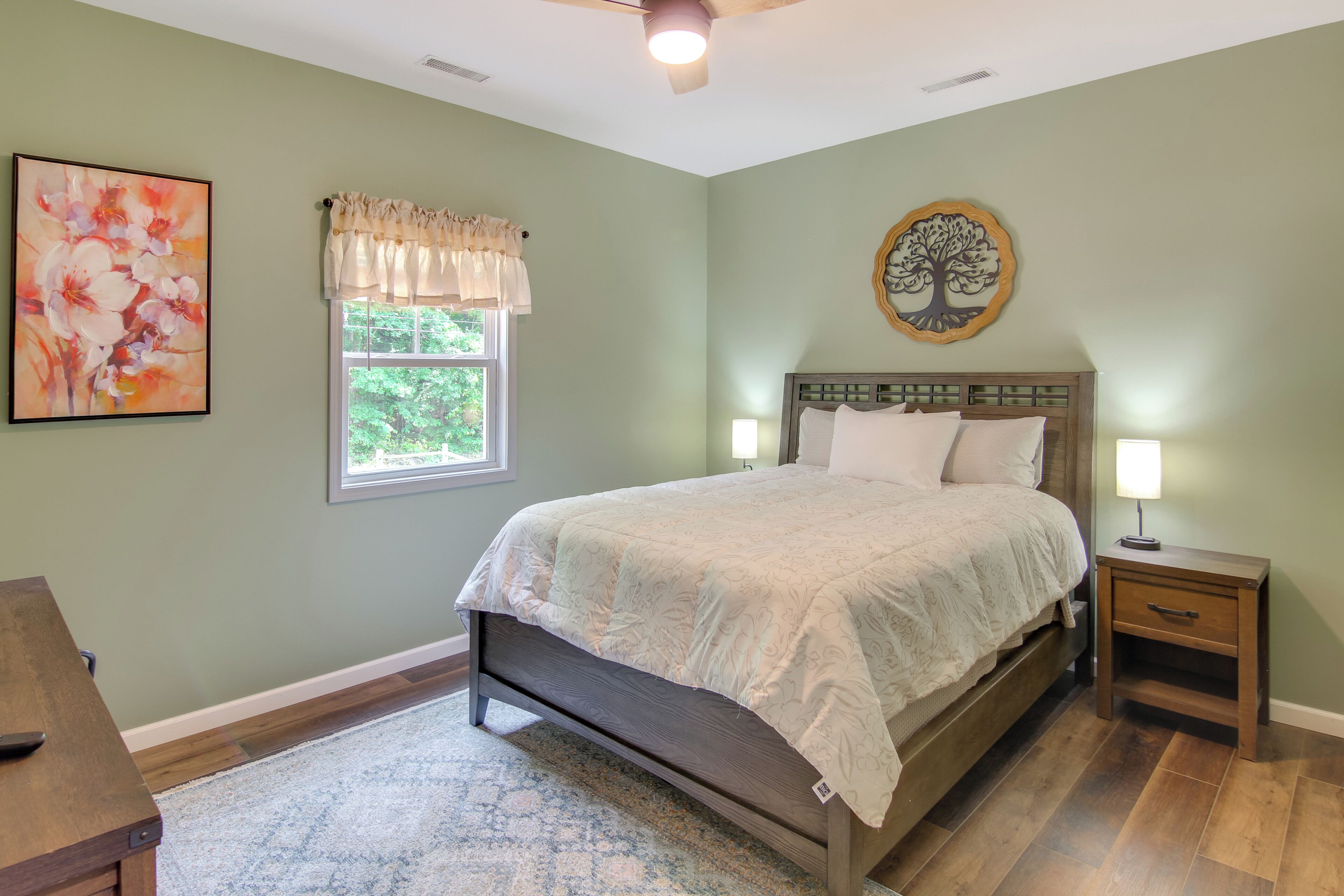 A bedroom with a bed, bedside tables, a window with curtains, a wall painting, and a decorative tree wall art.