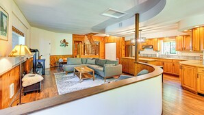 A spacious living area with a curved kitchen, wooden flooring, and a large window providing natural light. - 5 Mi to Mt Rainier Natl Park: Cozy Cabin w/ Deck! (Ashford)