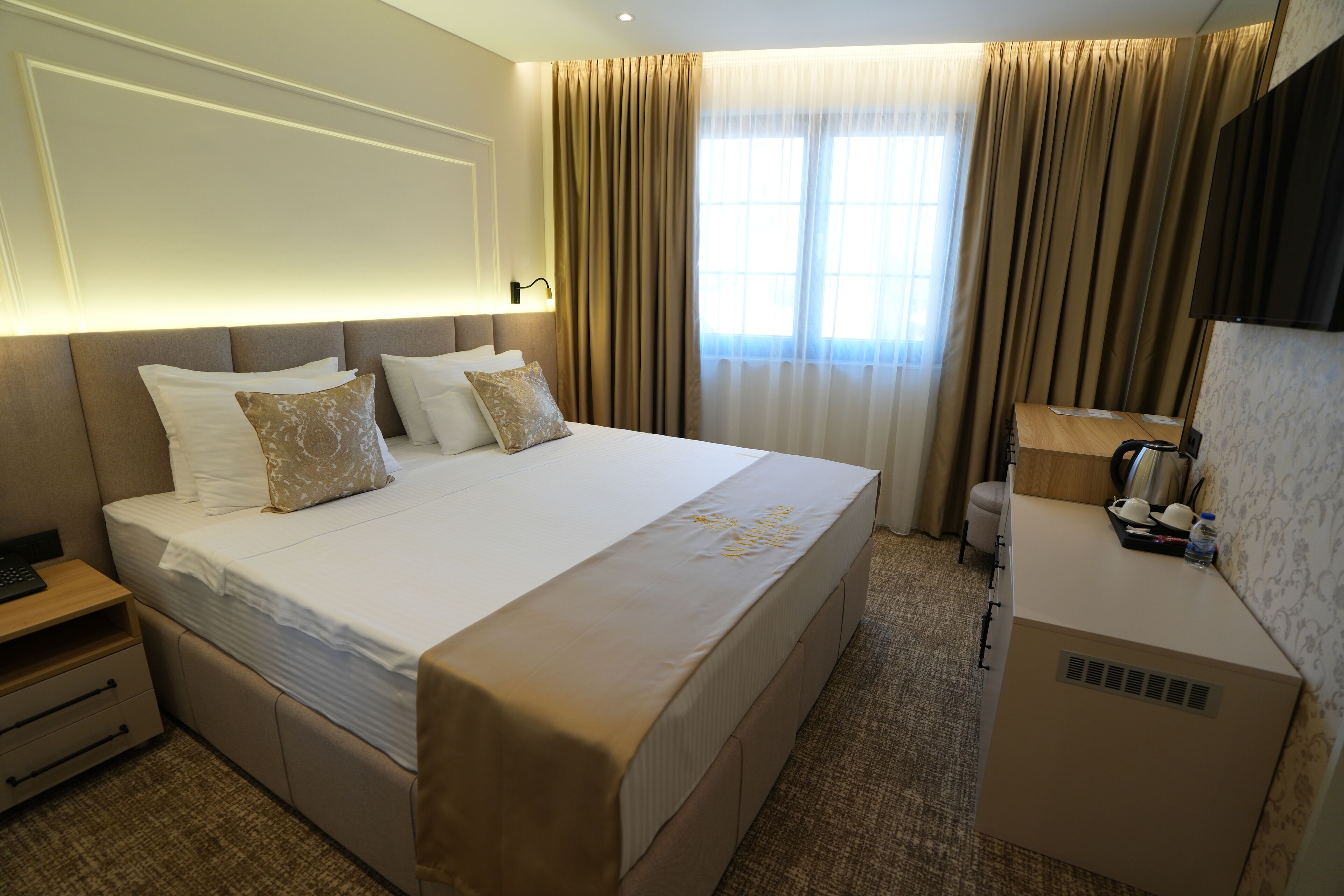 A hotel room with a large bed, bedside tables, a desk with a chair, a television, and a window with curtains.